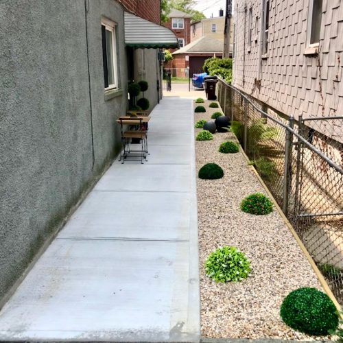 A serene walkway lined with lush plants, featuring a wooden bench inviting visitors to relax and enjoy the surroundings.