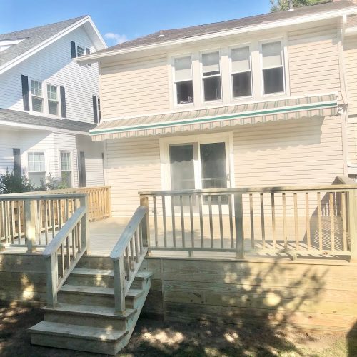 A wooden deck with stairs leading up to a charming house, surrounded by greenery and sunlight.